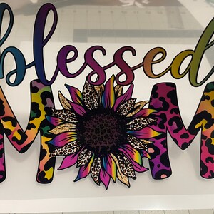 Peut inclure: Un autocollant en vinyle color&eacute; avec les mots "blessed mom" dans un d&eacute;grad&eacute; arc-en-ciel. Les lettres sont soulign&eacute;es de noir et ont un motif l&eacute;opard. Un grand tournesol avec un centre imprim&eacute; l&eacute;opard se trouve au milieu de l'autocollant.