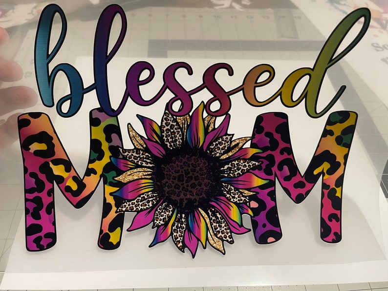 Peut inclure: Un autocollant en vinyle color&eacute; avec les mots "blessed mom" dans un d&eacute;grad&eacute; arc-en-ciel. Les lettres sont soulign&eacute;es de noir et ont un motif l&eacute;opard. Un grand tournesol avec un centre imprim&eacute; l&eacute;opard se trouve au milieu de l'autocollant.