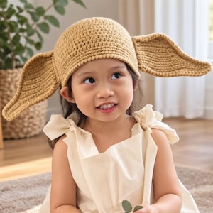 May include: A tan crochet hat featuring two large, pointed, protruding ears on either side. The hat is constructed with a dense, textured stitch pattern and is designed to cover the top of the head, extending outward with wide, flat, triangular ear flaps.