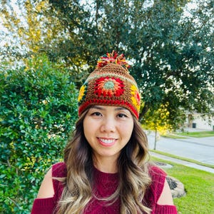 May include: A colorful, crocheted beanie hat with a pom-pom on top. The hat features a floral design in shades of brown, red, yellow, and green. The hat is worn by a person with long, wavy brown hair and a burgundy sweater.