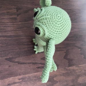 May include: A green crocheted plush toy with large black eyes and long arms. The toy is standing on a wooden surface.