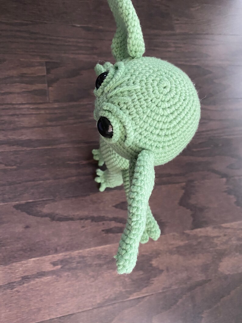 May include: A green crocheted plush toy with large black eyes and long arms. The toy is standing on a wooden surface.