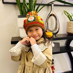 May include: A child wearing a brown crochet hat with colorful floral designs and a large pom-pom. The child is also wearing a tan coat with a white lace collar and white cuffs. The background includes a plant, a silver vase, and a decorative sign.