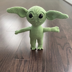 May include: Green crocheted plush toy resembling a character from the Star Wars franchise. The toy has large ears, big eyes, and a small mouth.