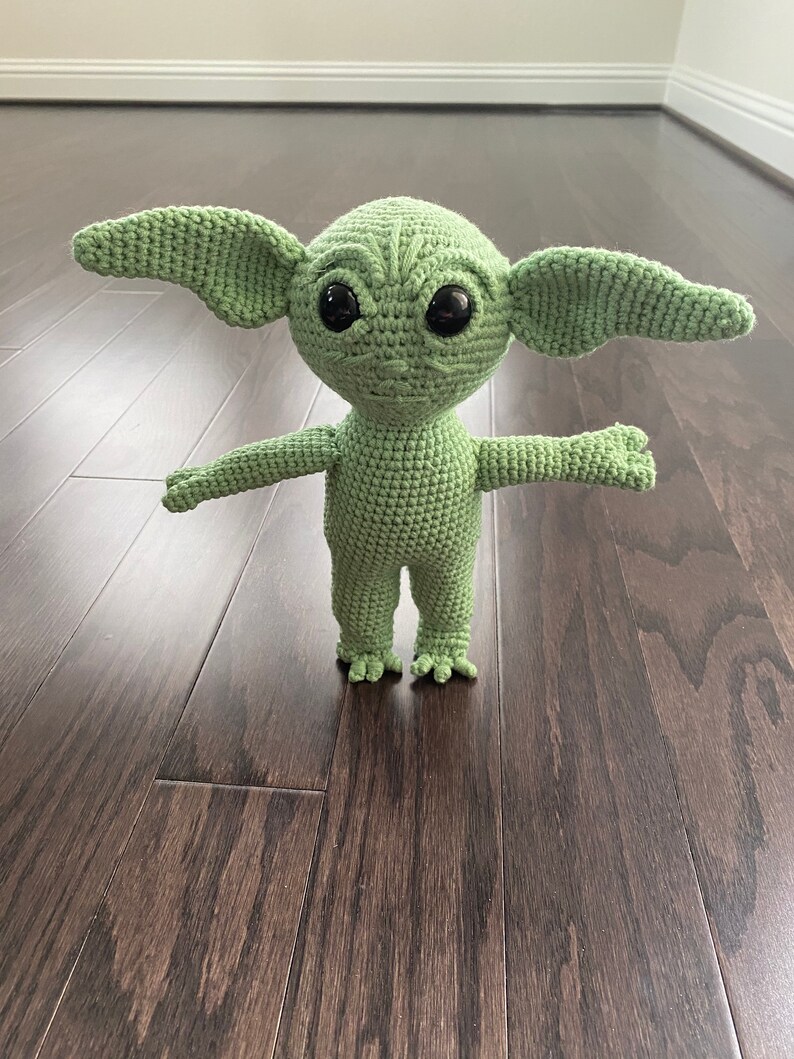 May include: Green crocheted plush toy resembling a character from the Star Wars franchise. The toy has large ears, big eyes, and a small mouth.