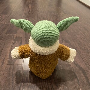 May include: A crocheted green and brown plush toy with large ears. The toy is wearing a brown fuzzy coat with white trim.