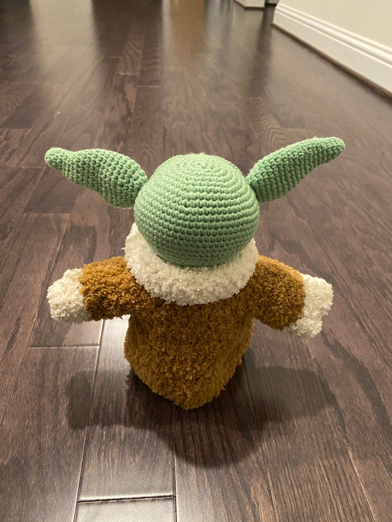May include: A crocheted green and brown plush toy with large ears. The toy is wearing a brown fuzzy coat with white trim.