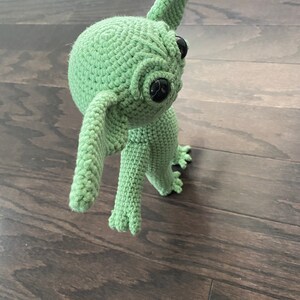 May include: A green crocheted plush toy with large black eyes and a long, pointed ear. The toy is standing on a wooden floor.
