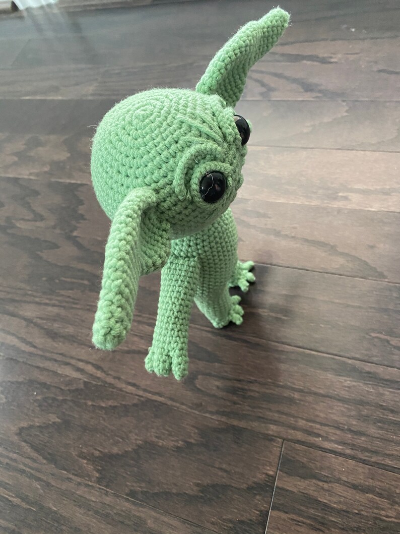 May include: A green crocheted plush toy with large black eyes and a long, pointed ear. The toy is standing on a wooden floor.
