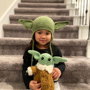 May include: A young child wearing a green crocheted Yoda hat with large ears, holding a green crocheted Yoda plush toy. The child is sitting on a set of stairs.