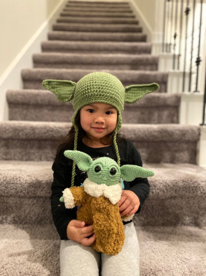 May include: A young child wearing a green crocheted Yoda hat with large ears, holding a green crocheted Yoda plush toy. The child is sitting on a set of stairs.