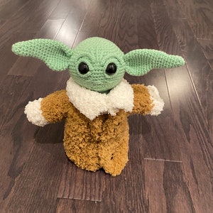 May include: A green crocheted plush toy with large eyes and a brown fuzzy coat. The toy is standing on a dark wood floor.
