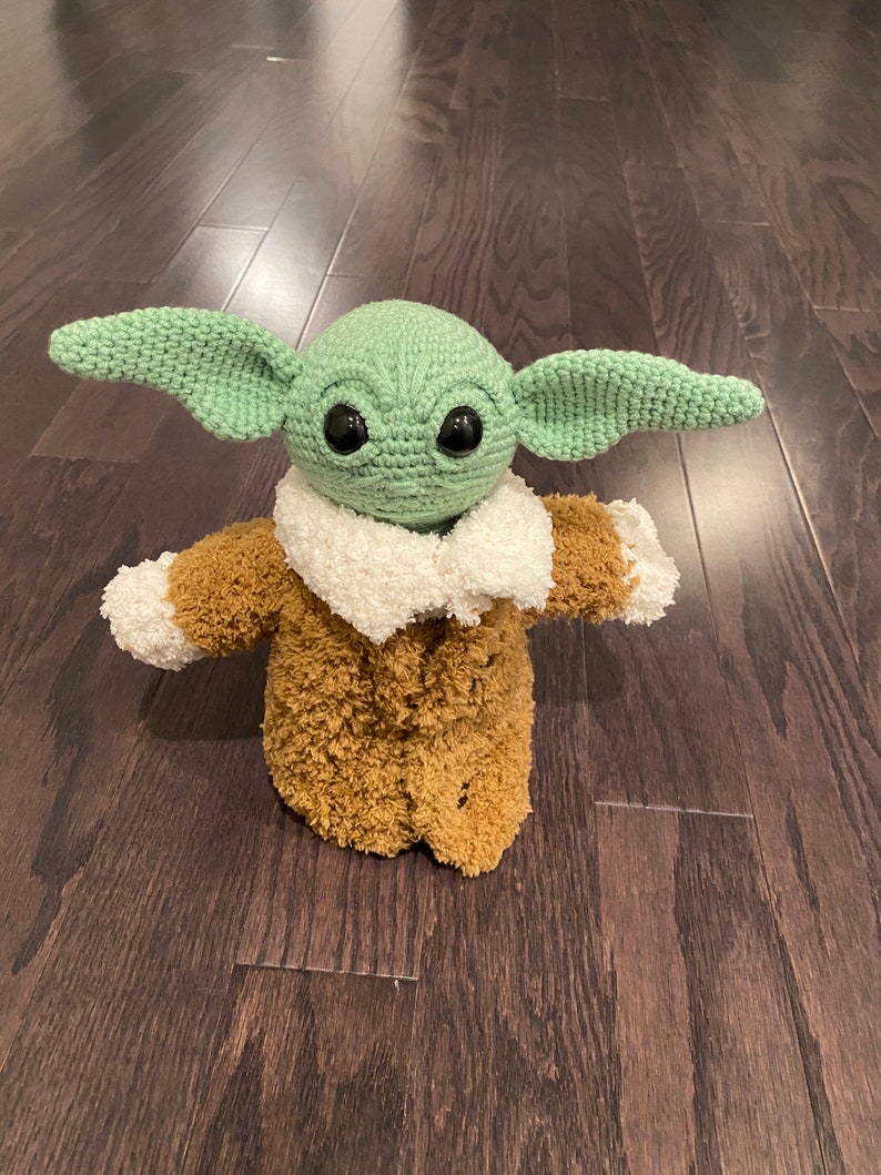 May include: A green crocheted plush toy with large eyes and a brown fuzzy coat. The toy is standing on a dark wood floor.