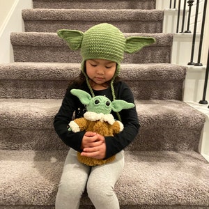 May include: A young person wearing a green crocheted hat with ears sits on a carpeted staircase holding a green crocheted plush toy.