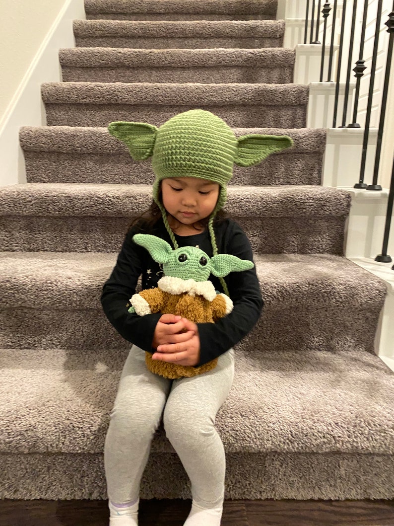 May include: A young person wearing a green crocheted hat with ears sits on a carpeted staircase holding a green crocheted plush toy.