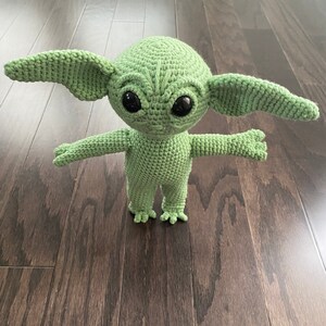 May include: Green crocheted plush toy resembling a character from the Star Wars franchise. The toy has large ears, big eyes, and a small mouth. It is standing on a wooden floor.