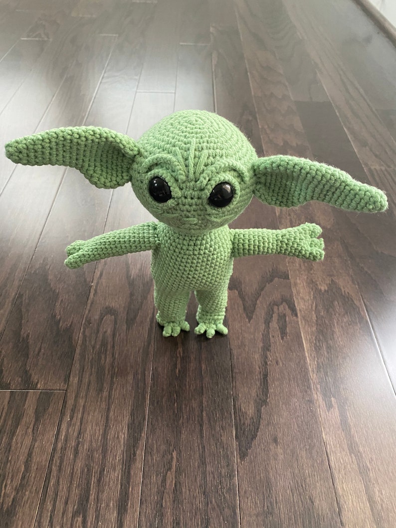 May include: Green crocheted plush toy resembling a character from the Star Wars franchise. The toy has large ears, big eyes, and a small mouth. It is standing on a wooden floor.
