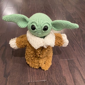 May include: A green crocheted plush toy with large eyes and a brown fuzzy coat. The toy is standing on a wooden floor.