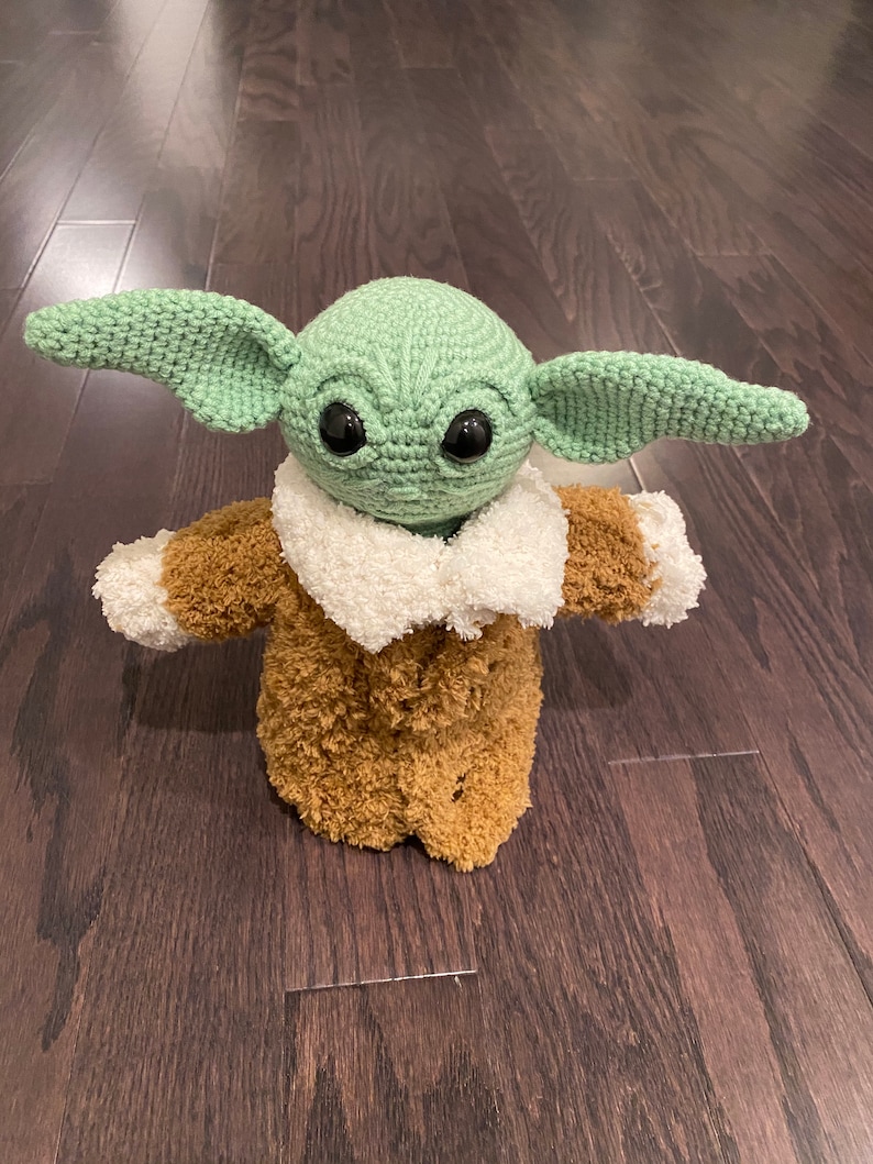 May include: A green crocheted plush toy with large eyes and a brown fuzzy coat. The toy is standing on a wooden floor.