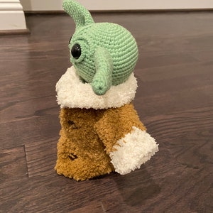 May include: A green crocheted plush toy with large eyes and a long nose, wearing a brown fuzzy coat with white trim.