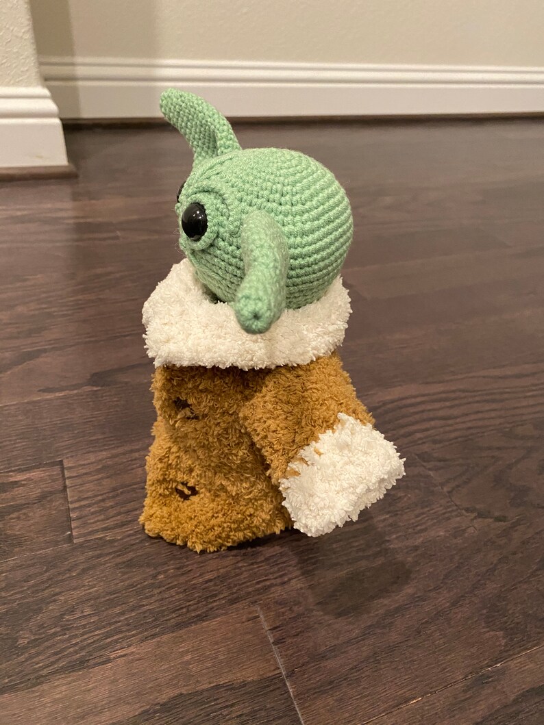 May include: A green crocheted plush toy with large eyes and a long nose, wearing a brown fuzzy coat with white trim.