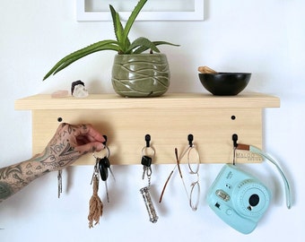 May include: A light wood key rack with five hooks, a small potted plant, a black bowl, and a light blue instant camera with the text "MALOO DESIGN" on the strap. Keys, glasses, and a small silver tube are hanging from the hooks.