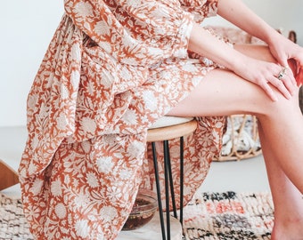 May include: A woman wearing a long, flowing, rust-colored dress with a white floral pattern. The dress has a gathered skirt and long sleeves. She is sitting on a stool with her legs crossed.