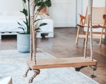May include: A wooden swing hanging from the ceiling in a room with a light wood floor and a rug. The swing is made of natural wood and has thick rope.