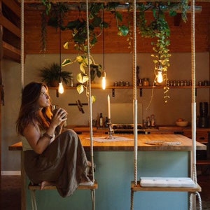 May include: Two wooden swing seats suspended by thick ropes from a ceiling beam, positioned in front of a kitchen island with a light blue base and a wooden countertop. Hanging green plants and three pendant light bulbs hang above the island.
