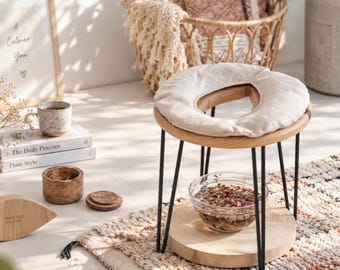 May include: A wooden yoni steam seat with a round light-coloured fabric cushion, supported by four thin black metal hairpin legs. The seat is positioned over a circular wooden base holding a clear glass bowl filled with dried herbs and flower petals.