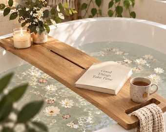 May include: A rectangular wooden bathtub tray resting across a white bathtub filled with water and floating white flowers. The tray holds a lit candle in a glass jar, a book with the text Good Things Take Time, and a speckled ceramic mug.