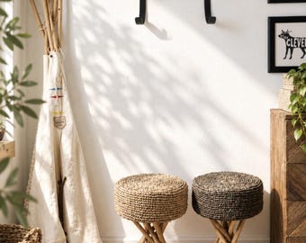 May include: Two round stools with woven seats and tripod-style wooden legs. One seat is light tan, the other is dark brown. The stools are positioned in front of a white wall with a black metal wall-mounted hook and a white fabric teepee nearby.