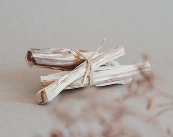May include: A bundle of brown and white palo santo sticks tied together with twine. The sticks are on a light brown surface.