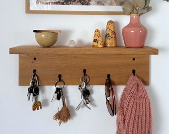 May include: A wooden wall-mounted key rack with five hooks. The rack is made of light brown wood and has a shelf on top. There are keys, a hat, and sunglasses hanging from the hooks.