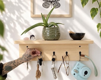 May include: A light-coloured wooden wall shelf featuring four black metal hooks. The shelf holds a green textured planter with an aloe plant, a small black bowl, and two crystals. Hanging from the hooks are keys, glasses, and a light blue camera.