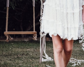 May include: A wooden swing set with rope and a white wooden frame. The swing is hanging from a tree branch. A person wearing a white dress is walking in the grass.
