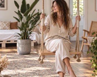 May include: A wooden indoor swing suspended by thick, natural-coloured twisted ropes. The swing features a rectangular wooden seat and two vertical rope sections with light-coloured fabric grips at the top, designed for interior use in a home setting.