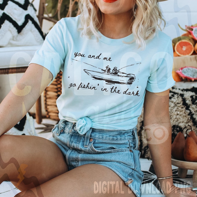 Puede incluir: Camiseta azul claro con el texto "you and me go fishin' in the dark" y una ilustraci&oacute;n en negro de un barco de pesca con dos personas. La camiseta est&aacute; anudada a la cintura y se combina con pantalones cortos vaqueros.