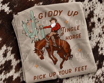 Puede incluir: Una camiseta beige con un gráfico de Santa Claus con sombrero de vaquero montando un caballo marrón encabritado junto a un cactus verde decorado con luces navideñas de colores. El texto dice GIDDY UP JINGLE HORSE PICK UP YOUR FEET.