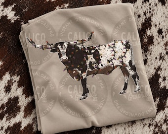 Pode incluir: Uma t-shirt bege claro com uma ilustração gráfica de uma vaca longhorn com manchas pretas e brancas, envolvida por um fio de luzes de Natal coloridas. O texto Calico Graphics é visível no centro do design.