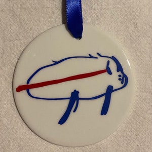 May include: A white ceramic ornament with a blue pig illustration and a red line across the middle.