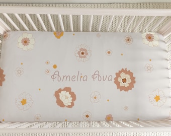 Peut inclure: Un lit bébé blanc avec un drap housse à motif floral dans les tons de brun, jaune et blanc. Le drap est brodé des noms "Amelia Ava" en brun.