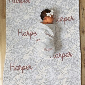 May include: A white baby blanket with a blue and white wave pattern. The blanket has the name "Harper" written on it multiple times in a brown cursive font.