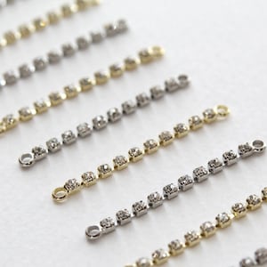 Può includere: Una collezione di catene d'argento e d'oro con strass quadrati. Le catene sono disposte in fila su una superficie bianca.