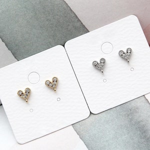 May include: Two pairs of heart-shaped earring charms displayed on white cards. One pair is gold-toned and the other is silver-toned. Each heart charm features a textured border and is embedded with small, clear, sparkling stones.