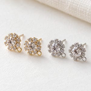 May include: Four stud earring components featuring a central round clear stone surrounded by a halo of smaller clear stones. Two components are gold-toned and two are silver-toned, each with a small circular loop at the bottom for attachments.