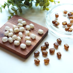 May include: A collection of tumbled gemstones in shades of white and brown. Some are on a small, square wooden board, while others are in a clear glass dish. The gemstones have a smooth, polished appearance and vary in size and color.