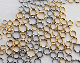 May include: An assortment of gold and silver-colored jump rings of various sizes. The rings are scattered across a white surface, with a mix of shiny gold and silver tones. These jewelry-making components are ideal for crafting.
