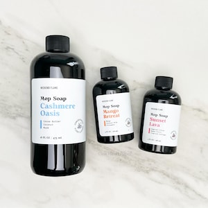 May include: Three black bottles of Weekend Flame Mop Soap. The largest bottle is labelled "Cashmere Oasis" with Cocoa Butter, Coconut, and Musk. The smaller bottles are "Mango Retreat" and "Sunset Lava". Each bottle has a black cap and a white label.