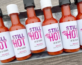 May include: Five small bottles of hot sauce with white labels that say "Still Hot" in pink and purple lettering. The labels also say "At 35 Years" and "Melissa's Bday Hot Sauce".